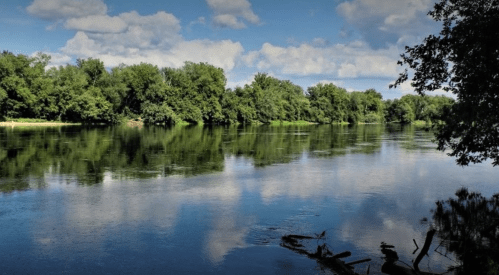 A serene river scene with lush green trees reflecting on calm water under a partly cloudy sky.