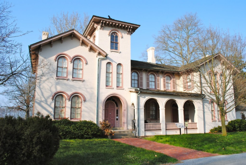 A large, historic two-story house with pink accents, arched windows, and a porch, surrounded by greenery.