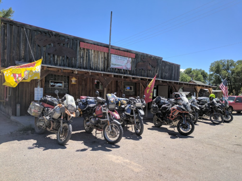 A row of motorcycles parked outside a rustic wooden building with flags and signage under a clear blue sky.