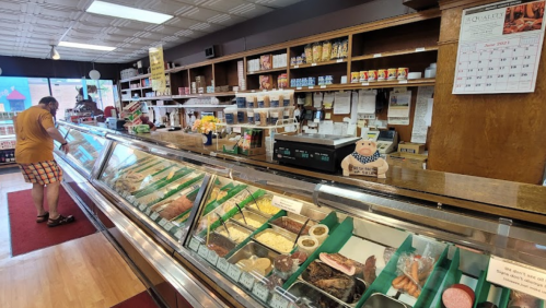 A deli display case filled with various meats and cheeses, with a customer browsing in the background.