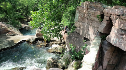A serene river flows through rocky terrain, surrounded by lush greenery and trees on a sunny day.