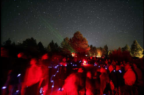 A crowd of people with glowing lights under a starry night sky, surrounded by trees and colorful laser lights.