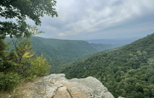 A rocky ledge overlooks a lush green valley with rolling hills under a cloudy sky.