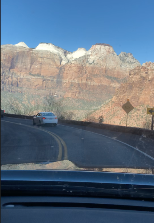 A winding road with a white car, surrounded by towering red rock mountains under a clear blue sky.