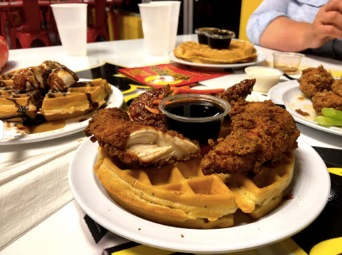 A plate featuring fried chicken on a waffle, served with syrup, alongside other dishes and drinks on a table.
