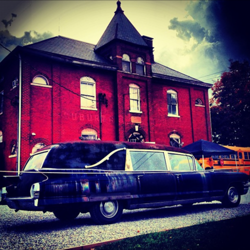 A vintage black hearse parked in front of a red brick building under a cloudy sky.