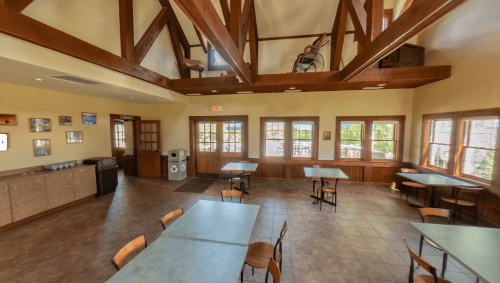A spacious, well-lit room with wooden beams, tables, and large windows, featuring a cozy, inviting atmosphere.
