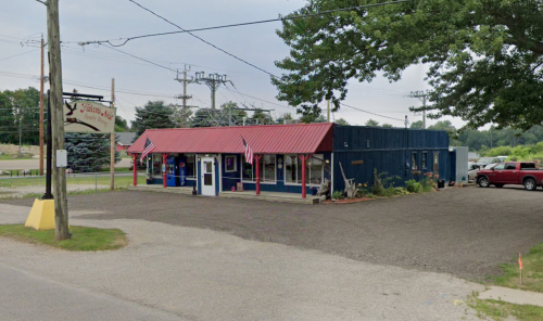 A small blue building with a red roof, flags, and a sign, located at a rural intersection with power lines nearby.