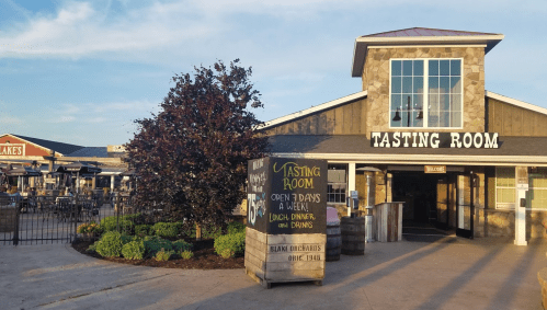 Exterior of a tasting room with a sign, surrounded by greenery and outdoor seating areas. Open daily for lunch and dinner.