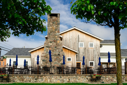 A rustic building with a stone chimney, surrounded by trees and outdoor seating with blue umbrellas.