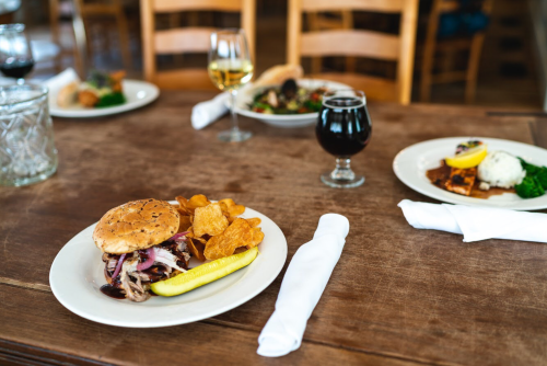 A plate with a pulled pork sandwich, chips, and a pickle, with drinks and other dishes in the background on a wooden table.
