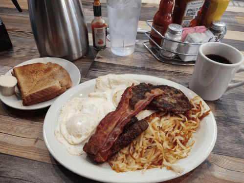 A plate of breakfast with eggs, bacon, sausage, hash browns, toast, and a cup of coffee on a wooden table.