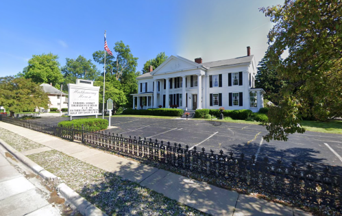 Historic white mansion with columns, surrounded by greenery and a black wrought-iron fence, on a sunny day.