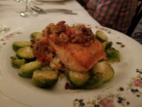 A plate of salmon topped with sautéed vegetables, served on a bed of Brussels sprouts. Elegant dining setting.