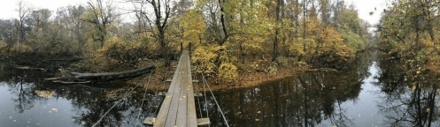 A wooden bridge spans a calm river surrounded by trees with autumn foliage in a serene landscape.