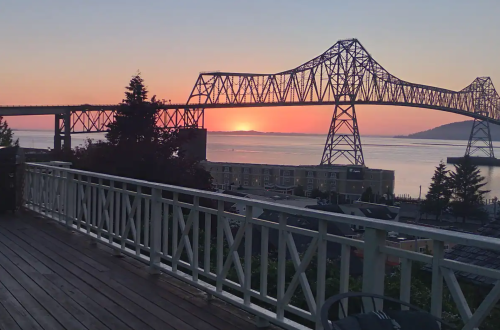 A sunset over a river, with a bridge silhouetted against the colorful sky, viewed from a wooden deck.