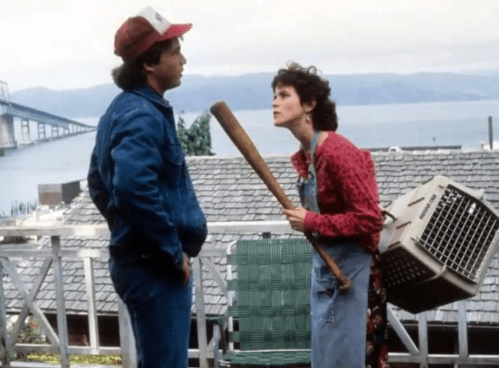 A man and woman stand outdoors, the woman holding a baseball bat and a pet carrier, with a bridge and water in the background.