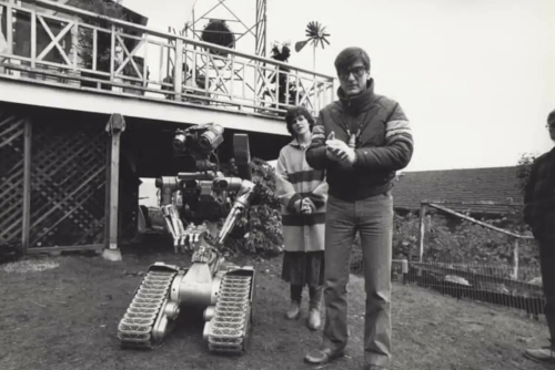 A man and a woman stand beside a robot on a grassy area, with a building and trees in the background.