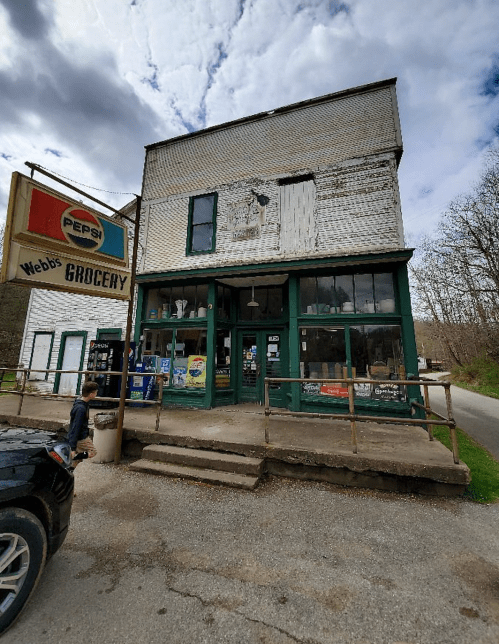 A vintage grocery store with a green facade and large windows, featuring a Pepsi sign outside.