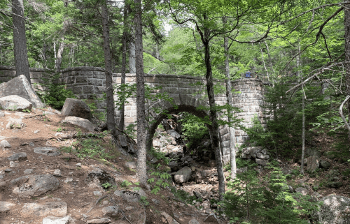 A stone bridge arches over a rocky creek, surrounded by lush green trees and a forested landscape.