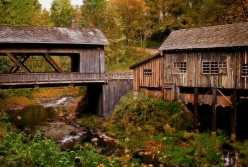 A rustic wooden covered bridge next to a weathered barn, surrounded by lush greenery and a flowing stream.