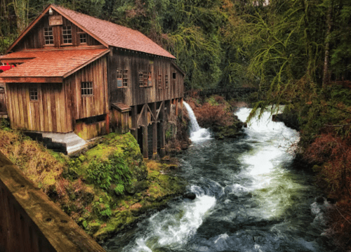 A rustic wooden mill beside a rushing river, surrounded by lush green trees and rocky terrain.