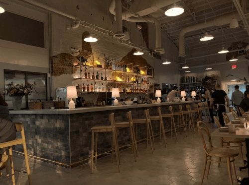 A modern restaurant interior featuring a bar with shelves of bottles, warm lighting, and wooden seating.