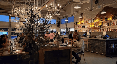 A stylish restaurant interior with warm lighting, patrons dining, and a bar stocked with various bottles.