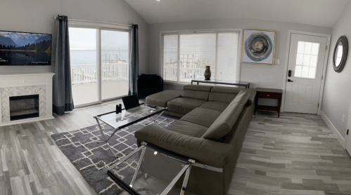 A modern living room with a sectional sofa, large windows, a TV, and a door leading to a deck with a scenic view.