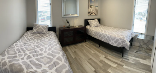 A bright bedroom with two twin beds, a nightstand, and large windows, featuring light wood flooring.