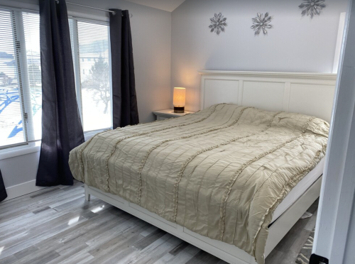 A cozy bedroom featuring a large bed with a beige comforter, a lamp, and windows with dark curtains.