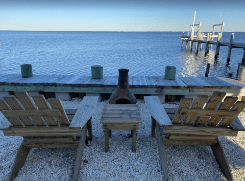 Two wooden chairs facing a calm water view, with a fire pit on a pebbled shore and a dock in the background.