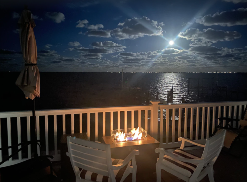 A serene night scene with a fire pit on a deck overlooking a calm waterway, illuminated by moonlight and clouds.