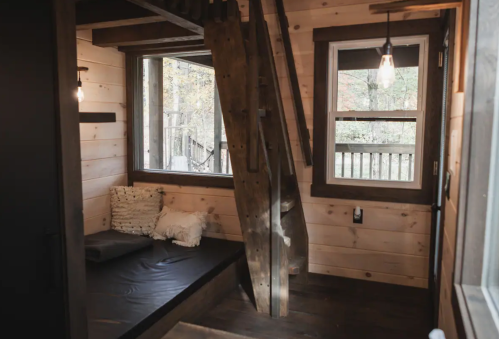 Cozy interior of a wooden cabin with a staircase, windows, and a small seating area. Natural light filters in.