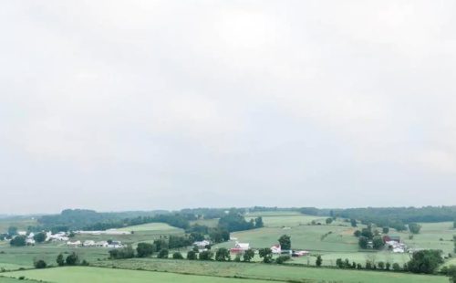 A serene landscape featuring rolling green hills and scattered farms under a cloudy sky.