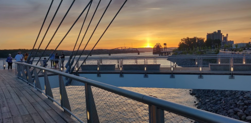 A sunset over a river, viewed from a bridge with people walking along the wooden path and city skyline in the background.