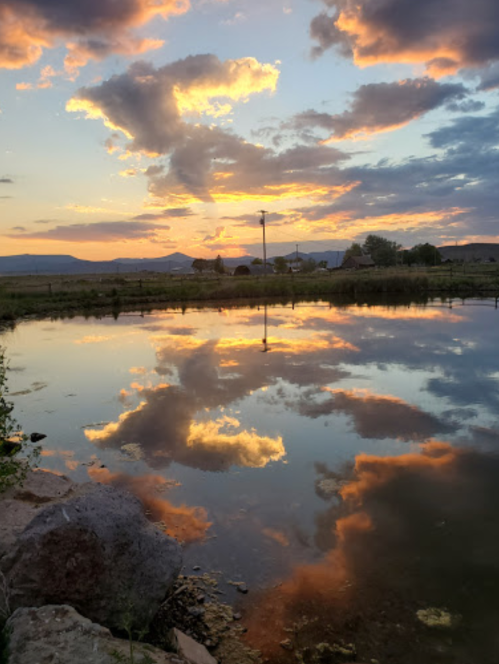 A serene sunset over a calm pond, with vibrant clouds reflecting in the water and mountains in the background.