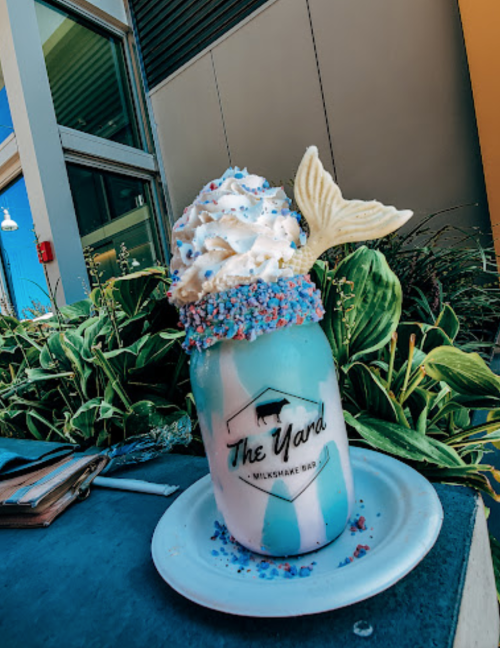 A colorful milkshake in a jar topped with whipped cream, sprinkles, and a mermaid tail, set against a leafy background.