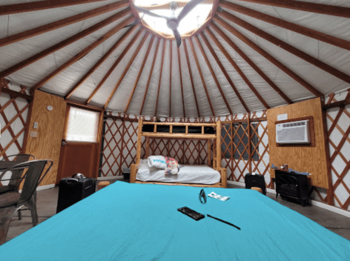 Interior of a yurt featuring a blue bedspread, a bunk bed, and wooden walls with a circular skylight.