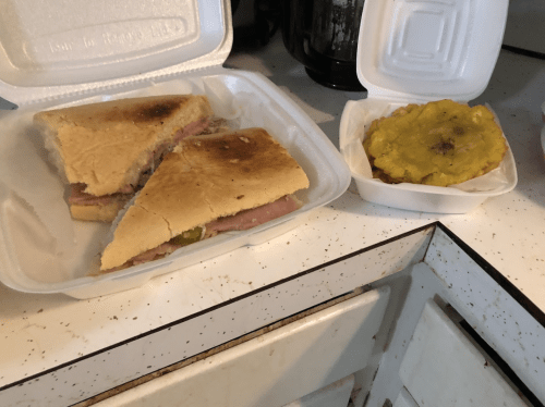 Two takeout containers: one with two ham sandwiches and the other with a fried green plantain.