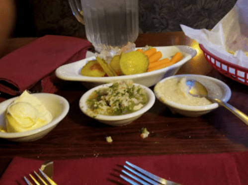 A table setting with bowls of pickles, carrots, coleslaw, and a side of butter, alongside utensils and a glass of water.