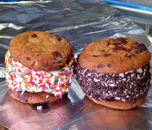 Two large cookie sandwiches filled with ice cream and coated in colorful sprinkles and chocolate sprinkles.