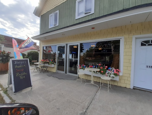 A cozy restaurant exterior with a sign for burgers, haddock, soup, and bubble tea, featuring flowers and outdoor seating.