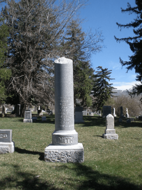 A tall, weathered gravestone in a cemetery, surrounded by trees and other headstones under a clear blue sky.