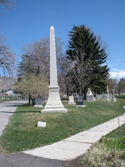 A tall white obelisk monument in a cemetery, surrounded by trees and gravestones under a clear blue sky.