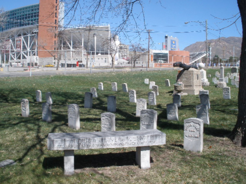 A cemetery with numerous gravestones, a bench, and a distant view of a stadium and mountains in the background.