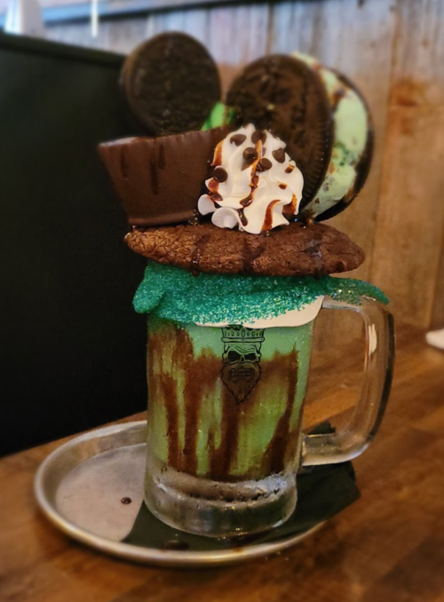 A tall glass filled with mint green dessert topped with cookies, whipped cream, and chocolate drizzle on a metal tray.