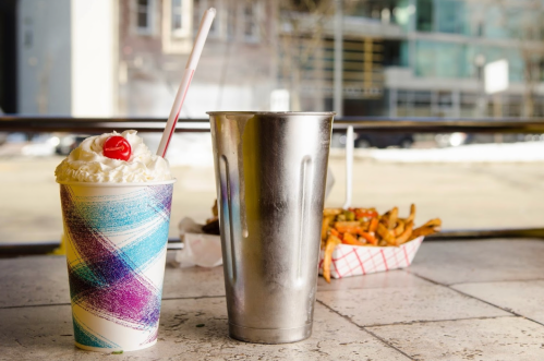 A colorful milkshake with whipped cream and a cherry sits next to a metal cup, with fries in the background.