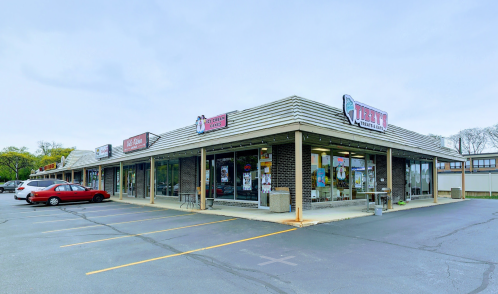 A strip mall with multiple storefronts, featuring a red car parked in front and a cloudy sky above.