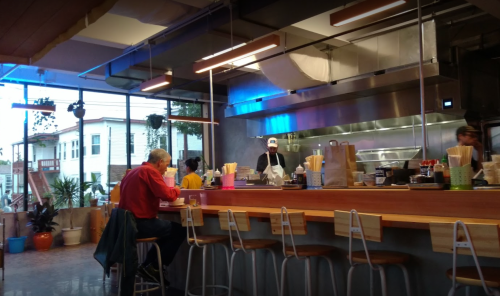 A modern restaurant interior with a counter, diners, and chefs preparing food in an open kitchen.
